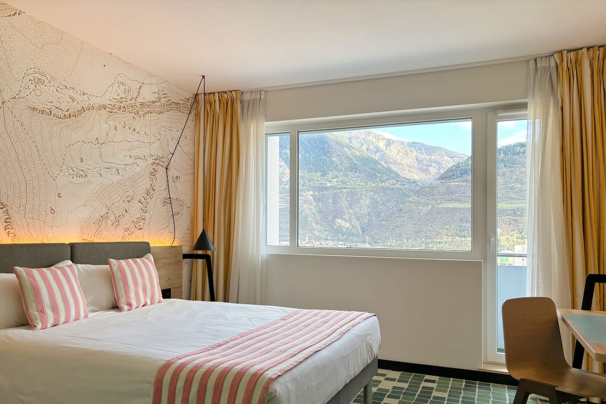 Habitación de hotel con una cama grande, un mapa en la pared, vistas de montañas a través de la ventana y una silla junto a la ventana.