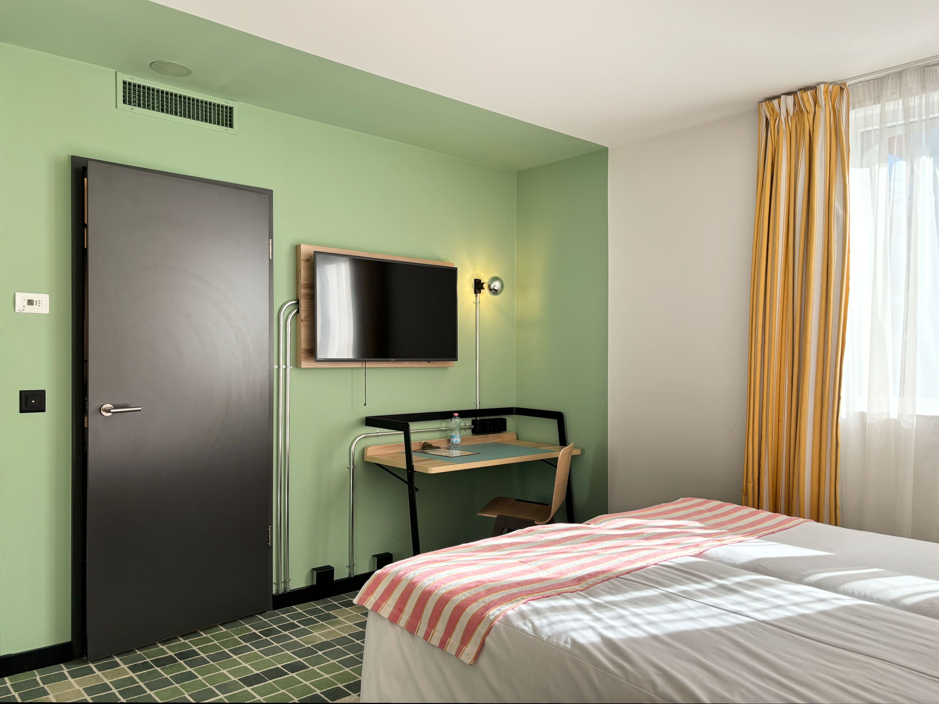 Una habitación de hotel con cama, escritorio con silla, televisor montado en la pared y una puerta.