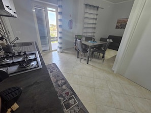 A kitchen with a gas stove, a dining table with chairs, a window with blinds, and a view of the outside.