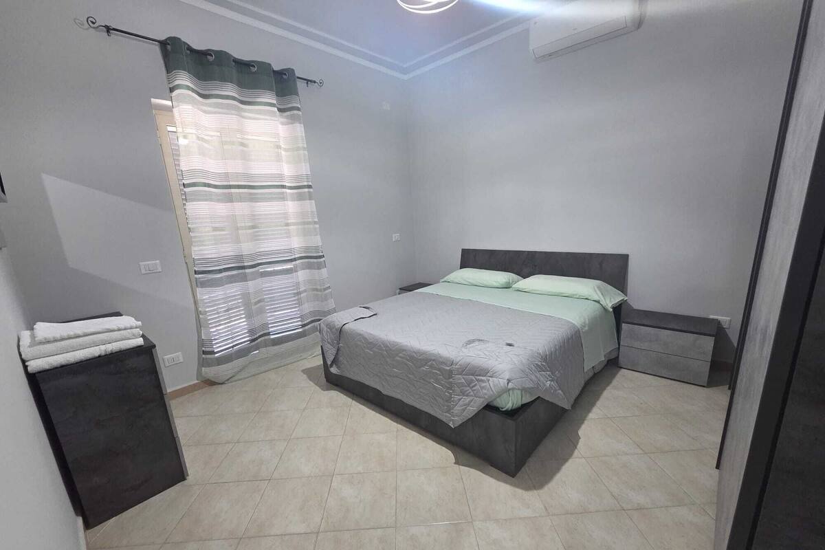 Une chambre à coucher avec un lit, une table de chevet, une petite armoire, un rideau et un luminaire au plafond.
