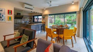 A modern living room with a sofa, dining table, and chairs. There is a kitchen area with a counter and cabinets. Large windows provide a view of the outdoors. - Cozy Nest 4Bhk Private Villa with Private Pool, Anjuna Beach North Goa (Vagator)