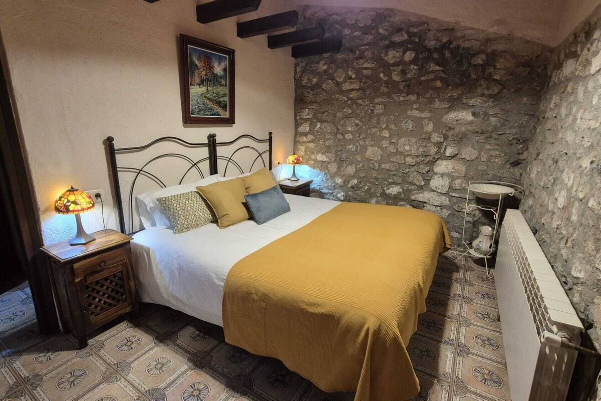 A bedroom with a stone wall, a bed with a yellow blanket, two bedside lamps, and a painting on the wall.