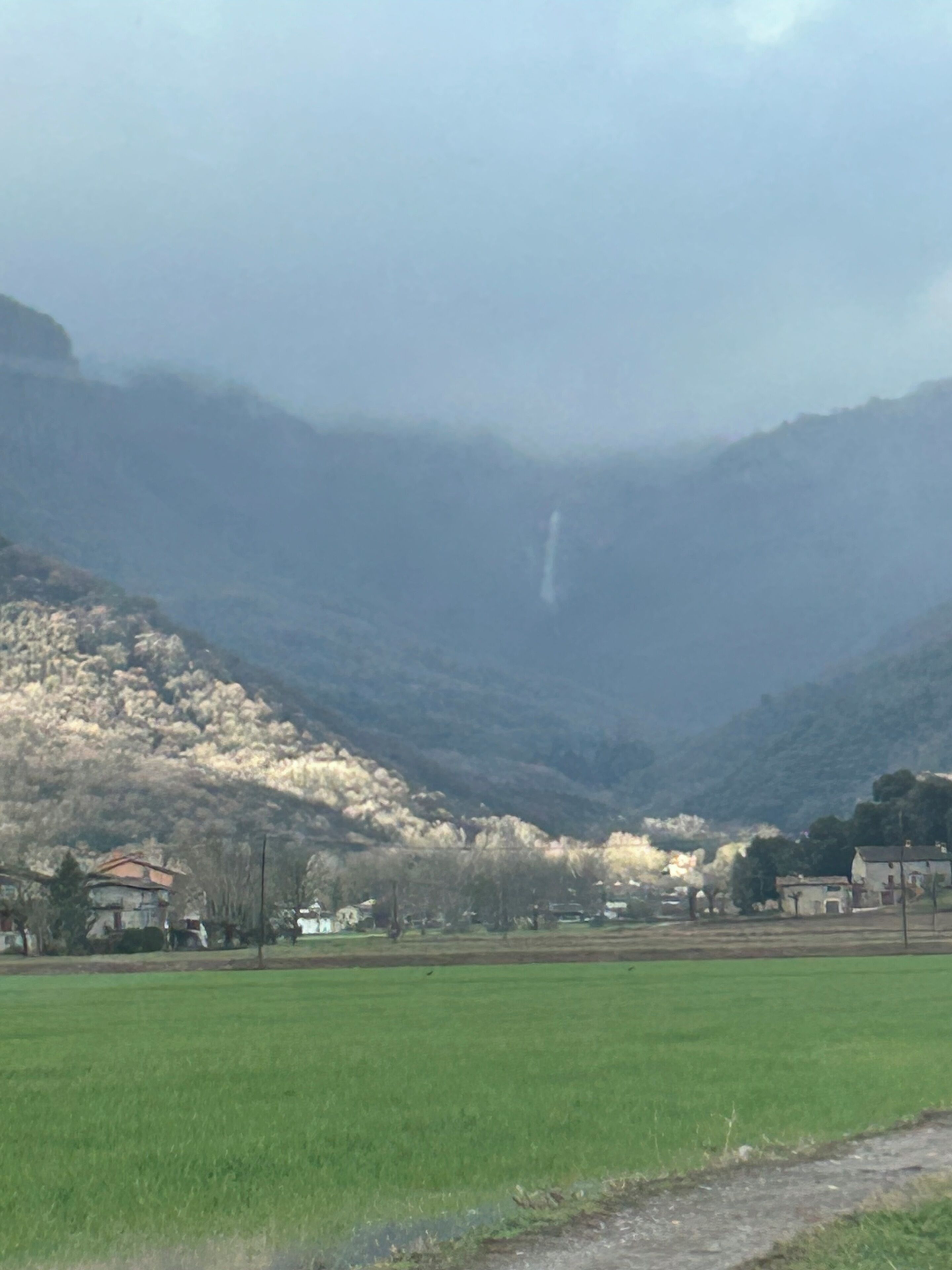 Paisaje rural con un campo verde, una montaña al fondo y una pequeña construcción con techo rojo.