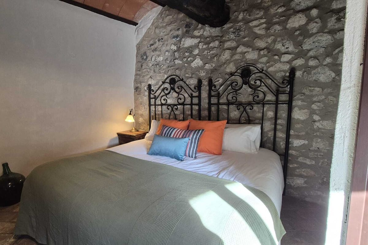 A bedroom with a stone wall, a metal bed frame, a bedside table with a lamp, and a green bedspread.