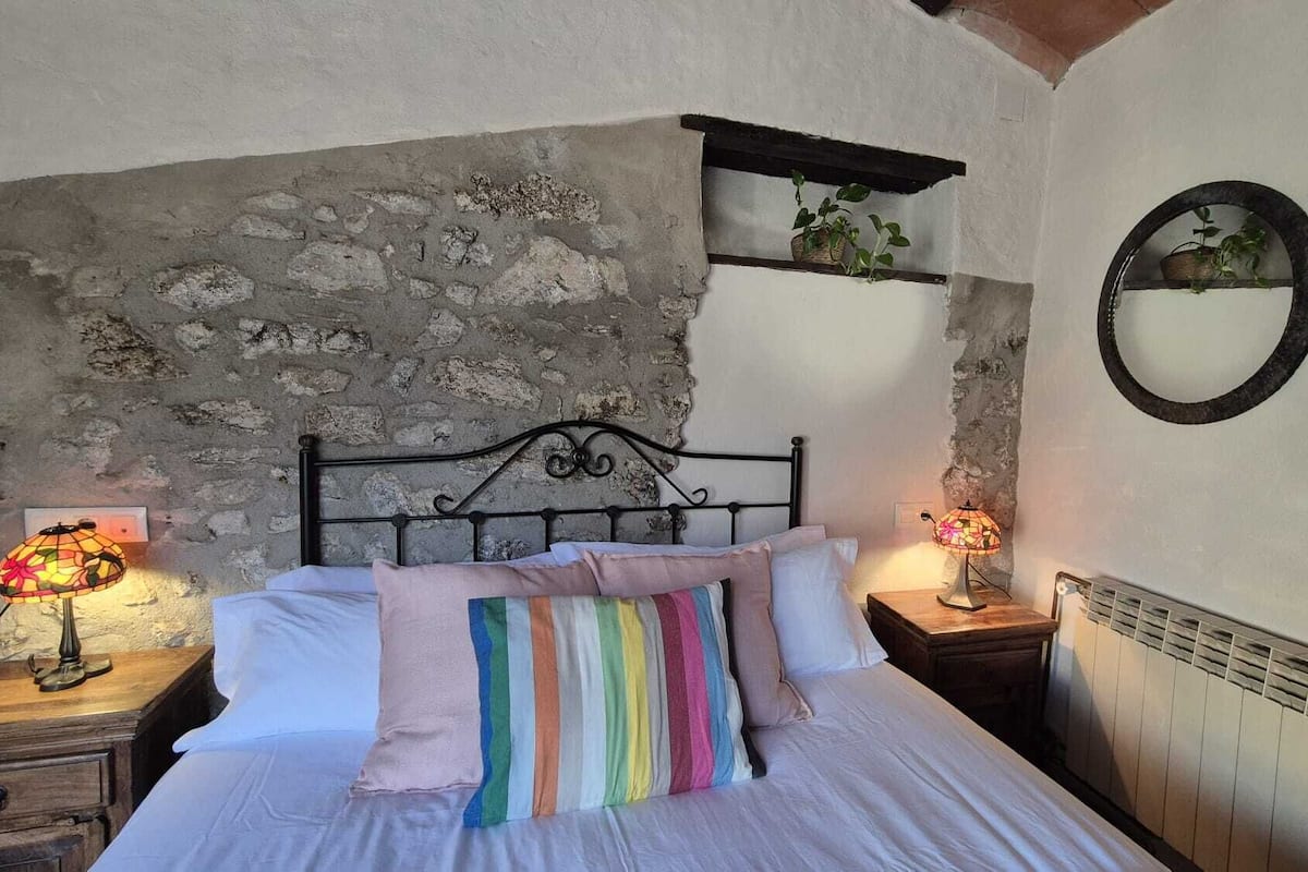 A bedroom with a stone wall, a bed with a rainbow-striped pillow, two bedside lamps, a wooden nightstand, a mirror, and a radiator.