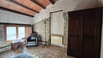Una habitación rústica con pared de piedra, puerta de madera, radiador, silla y ventana con contraventanas.
