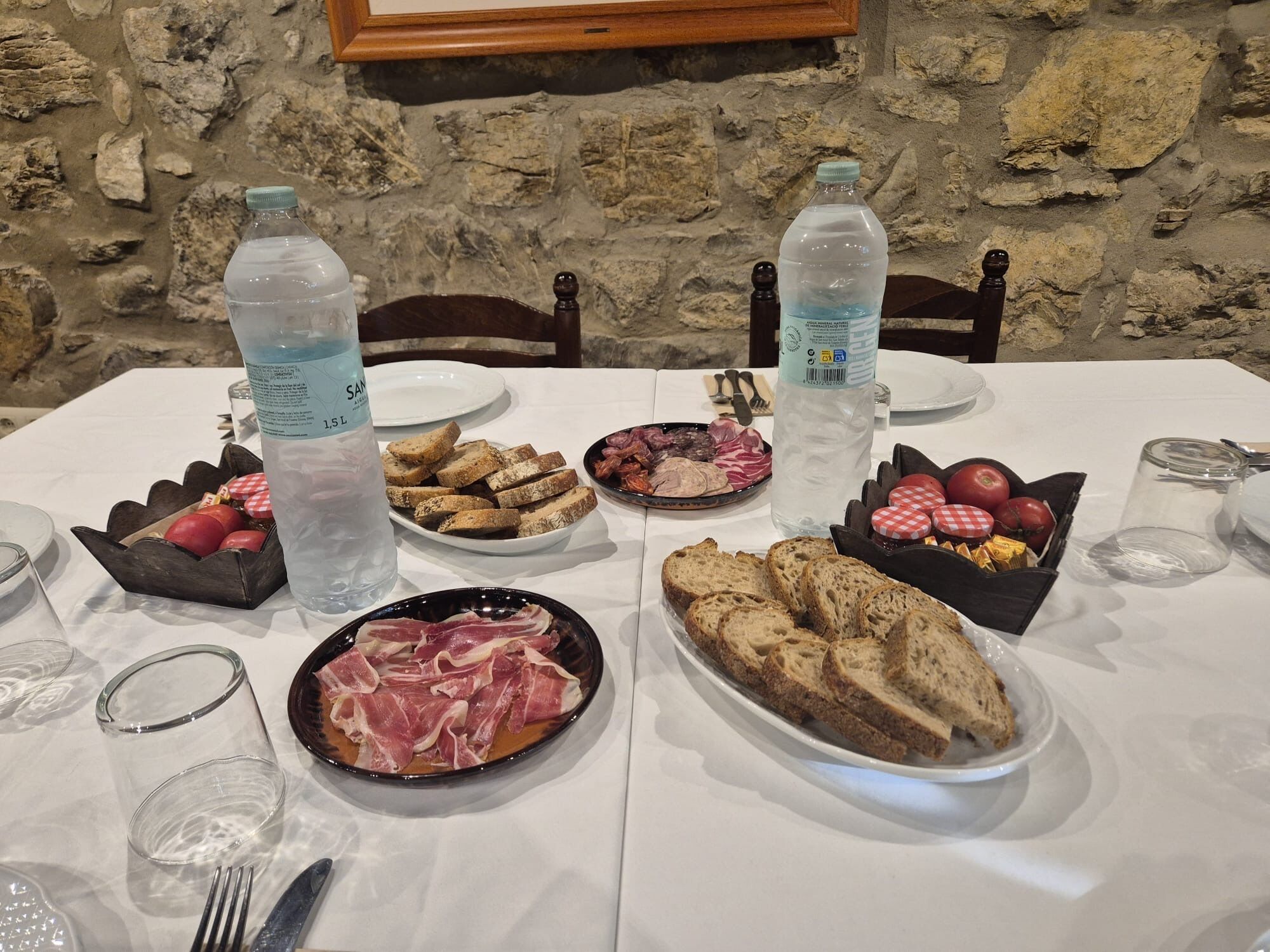 Una mesa dispuesta con diversos alimentos, como pan, carnes y frutas, junto con botellas de agua y cubiertos.