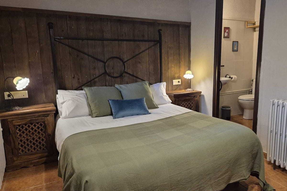 A bedroom with a wooden headboard, a bed with multiple pillows, bedside lamps, a radiator, and a view into the bathroom.
