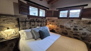 A bedroom with a stone wall, a bed with pillows, a nightstand with a lamp, and wooden windows. - Masia Rural Can Pei (Montagut)