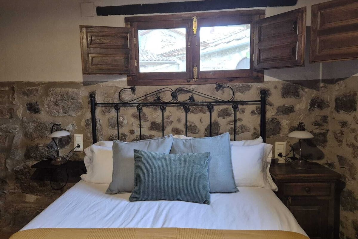 A bedroom with a stone wall, a bed with white linens, two bedside lamps, and a window with wooden shutters.