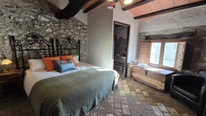 A rustic bedroom with a stone wall, a bed with colorful pillows, a wooden bench, and a black leather chair.