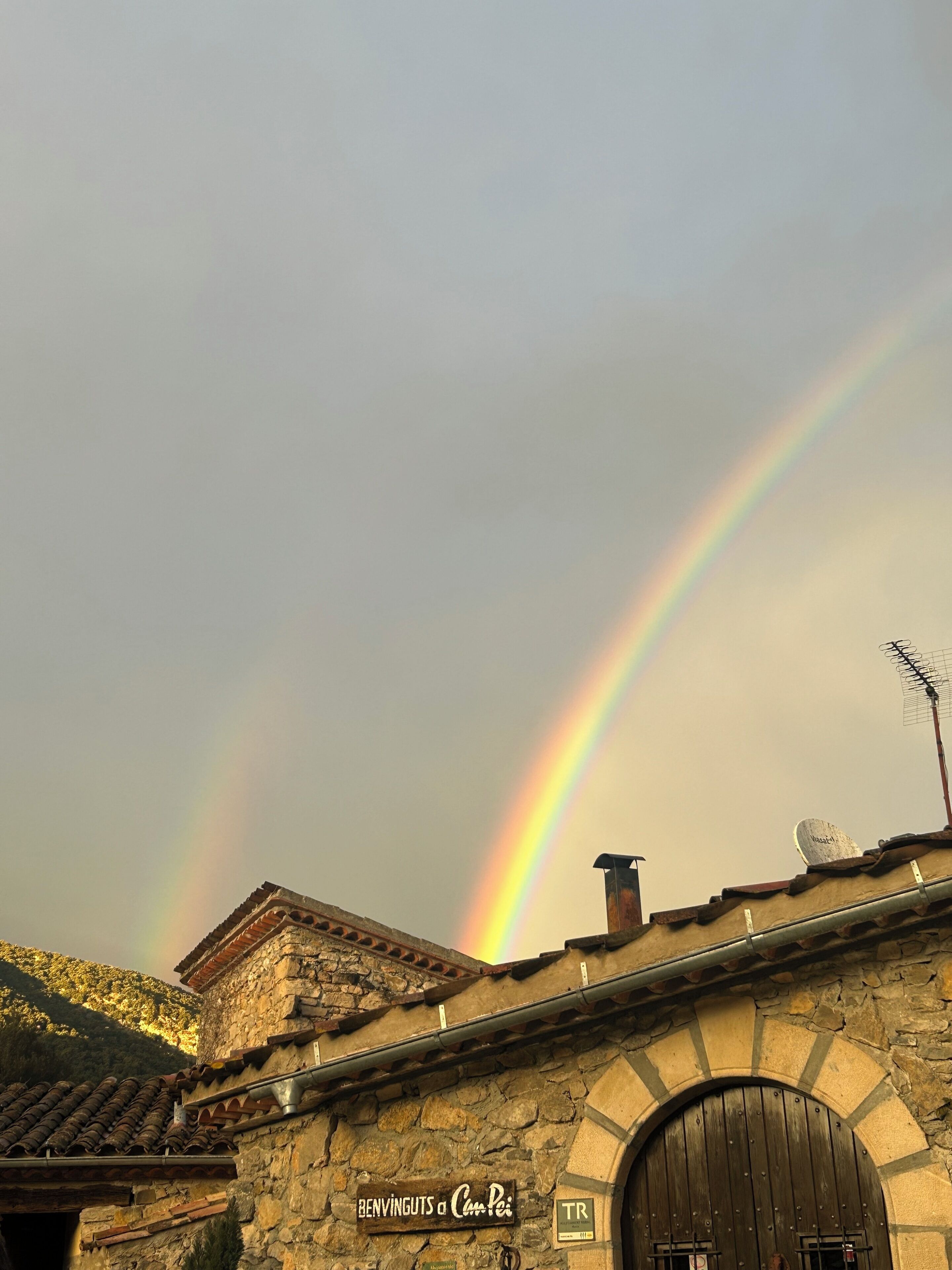 Un bâtiment en pierre avec une enseigne sur laquelle on peut lire « BENVINGUTS a Cam Pei » et un arc-en-ciel en arrière-plan.