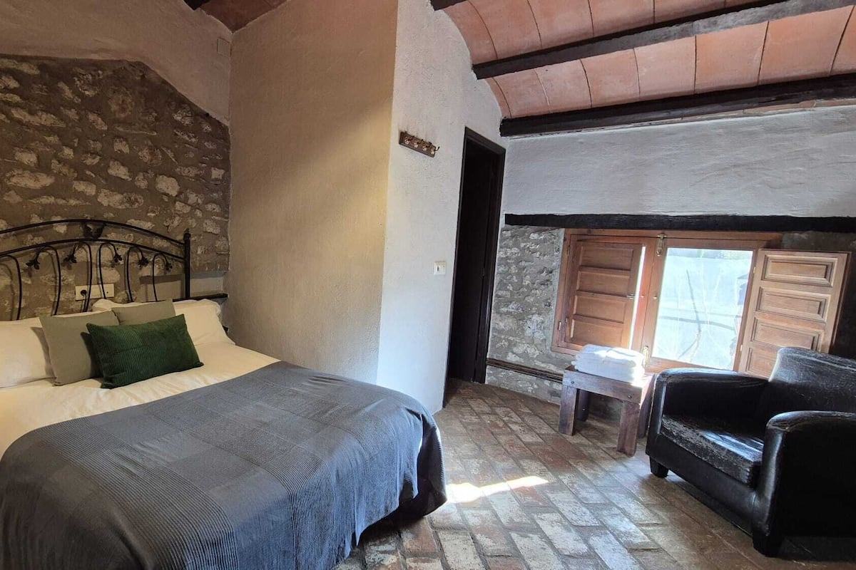 A bedroom with a stone wall, a bed with a headboard, a nightstand with a lamp, a wooden chair, and a small table with a towel.