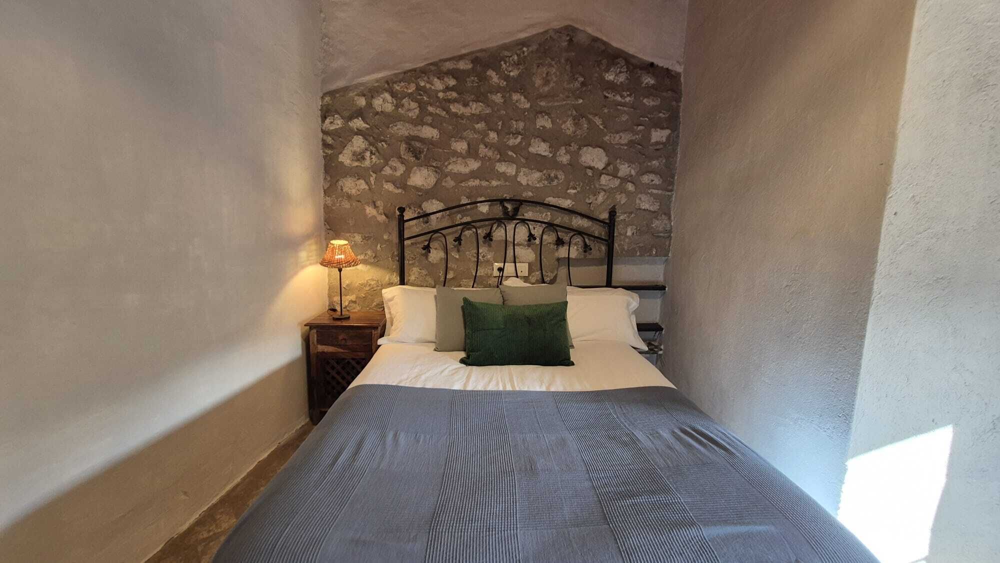 A narrow room with a stone wall, a bed with a metal headboard, a nightstand with a lamp, and a window allowing natural light.