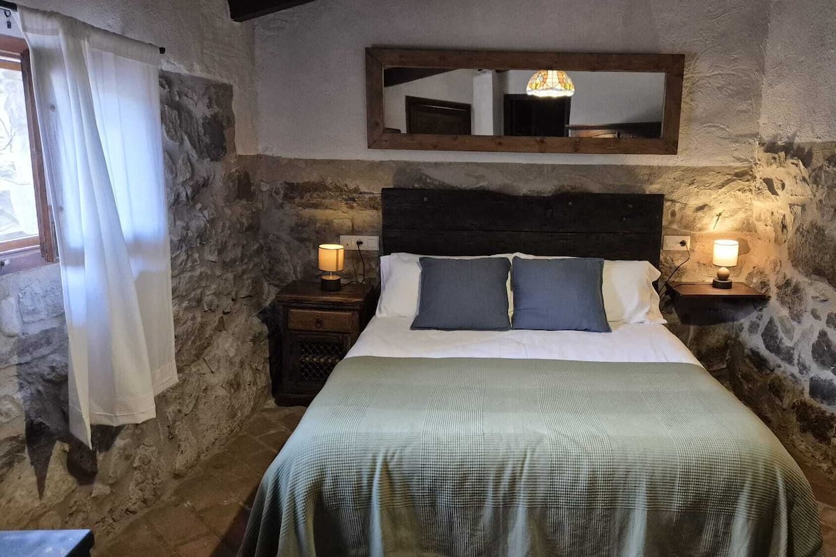 A stone-walled bedroom with a bed, two bedside tables with lamps, a mirror above the bed, and a window with curtains.