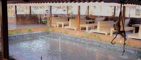 Un área de estar al aire libre con bancos y mesas de madera bajo un techo cubierto, con vista a una fuente de agua.