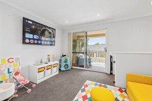 Una sala de estar moderna con un televisor de pantalla plana, un tapete de juegos colorido, un sofá amarillo y un área de juegos infantil con juguetes y espacio de almacenamiento.