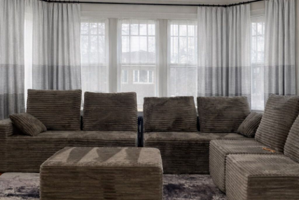 A living room with a sofa and ottoman, large windows with curtains, and a view of a building outside.