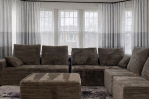 A living room with a sofa and ottoman, large windows with curtains, and a view of a building outside. - A 3-bedroom, 2-bathroom residence that provides ample space and comfort. (Chicago)