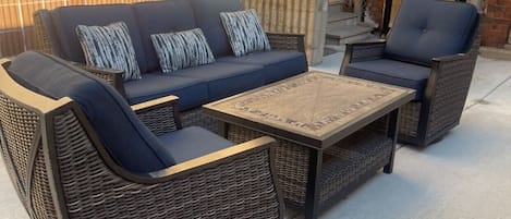 An outdoor patio with wicker furniture, including a sofa and chairs, and a wooden table.