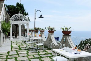 En udendørs terrasse med flisebelagte gulve, hvide metalstole, borde og plantekasser.