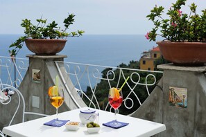 En terrasse med to plantekasser, et bord med hvid dug, to glas med drikkevarer, en lille skål med oliven og en dekorativ tallerken.
