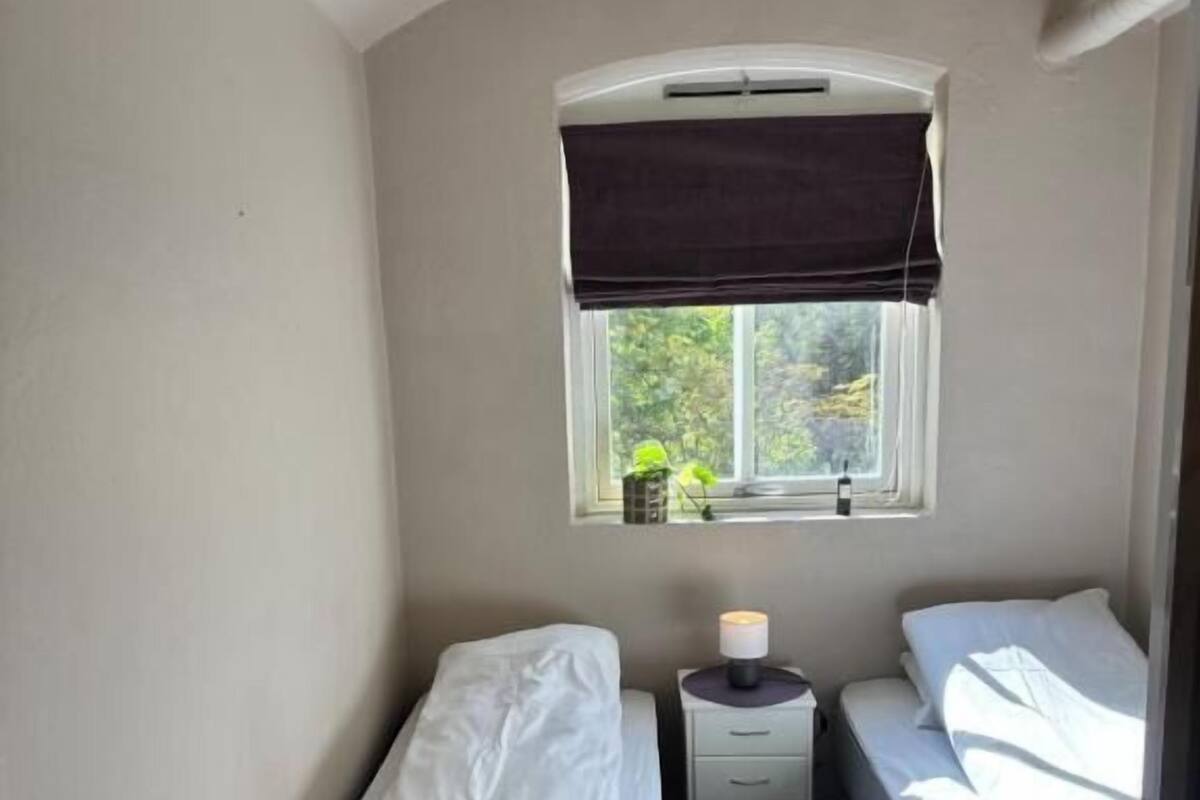 Un alojamiento pequeño de una sola habitación con dos camas, una ventana con vistas a la vegetación y una mesita de noche con lámpara.