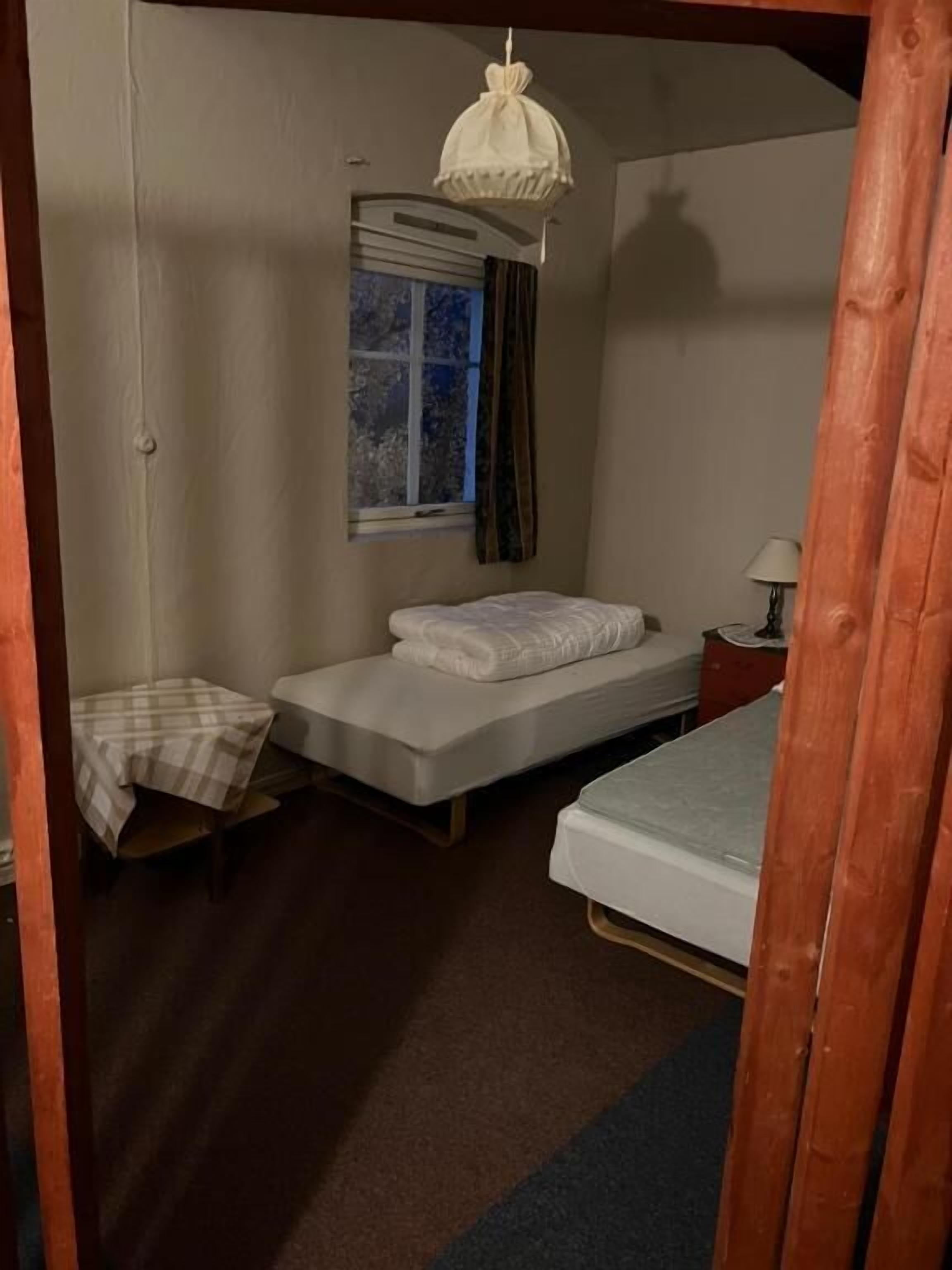 Une petite pièce, de taille modeste, comprenant deux lits simples, une petite table recouverte d’une nappe à carreaux, une fenêtre avec des rideaux et un luminaire suspendu.