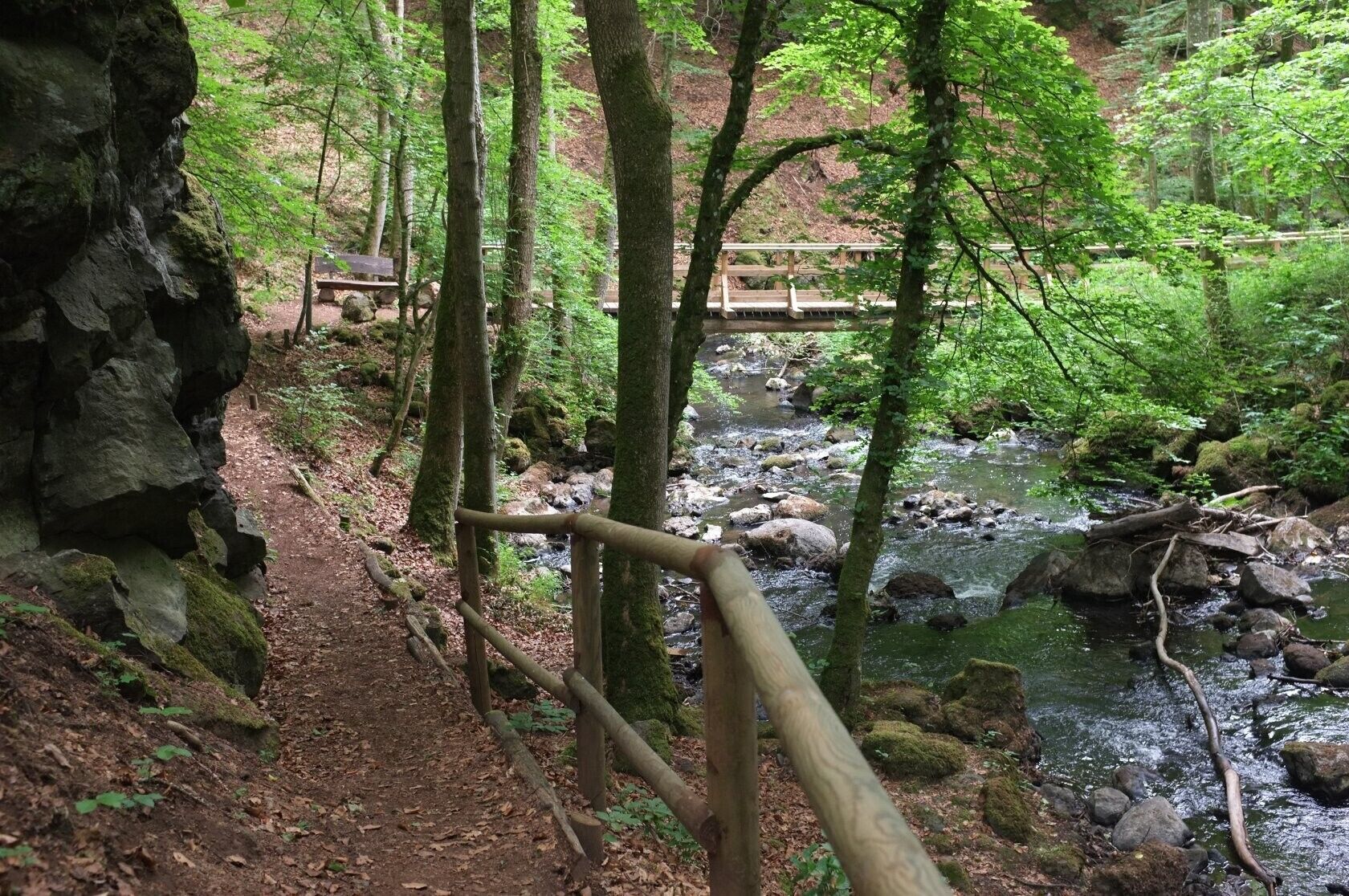 A forested area with a wooden bridge over a stream, a dirt path, and a rocky outcrop.