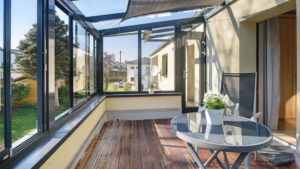 A sunlit balcony with a glass table, potted plants, and a view of neighboring houses.