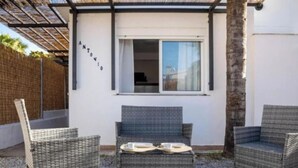 Een overdekt terras met rieten meubels en een tafel gedekt voor twee personen, grenzend aan een wit gebouw met een raam.