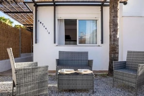A covered outdoor seating area with wicker furniture and a table set for two, adjacent to a white building with a window.