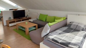 Una sala de estar moderna estilo loft con un televisor de pantalla plana, un sofá con cojines verdes, una mesa de centro y una cama con edredón a cuadros.