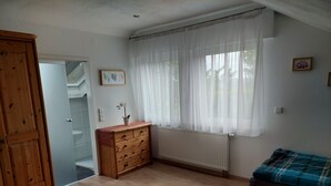 Una habitación con un aparador de madera, un radiador, una ventana con cortinas translúcidas, una cama con manta escocesa, y una puerta que da a otra habitación.