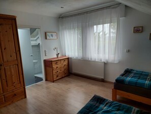 Une pièce comprenant une commode en bois, un radiateur, une fenêtre avec des rideaux légers, un lit recouvert d’une couverture à carreaux et une porte donnant sur une autre pièce.