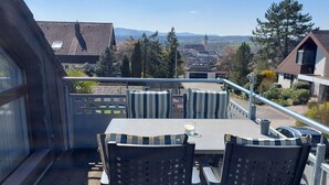 Una terraza en la azotea con mesa y sillas, baranda y vistas a edificios y árboles.
