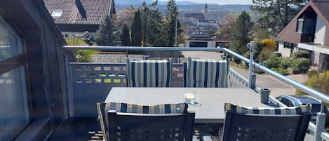 A rooftop terrace with a table and chairs, a railing, and a view of buildings and trees.