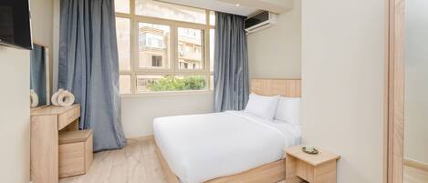 A bedroom with a bed, bedside table, a desk with drawers, a window with curtains, and an air conditioning unit.