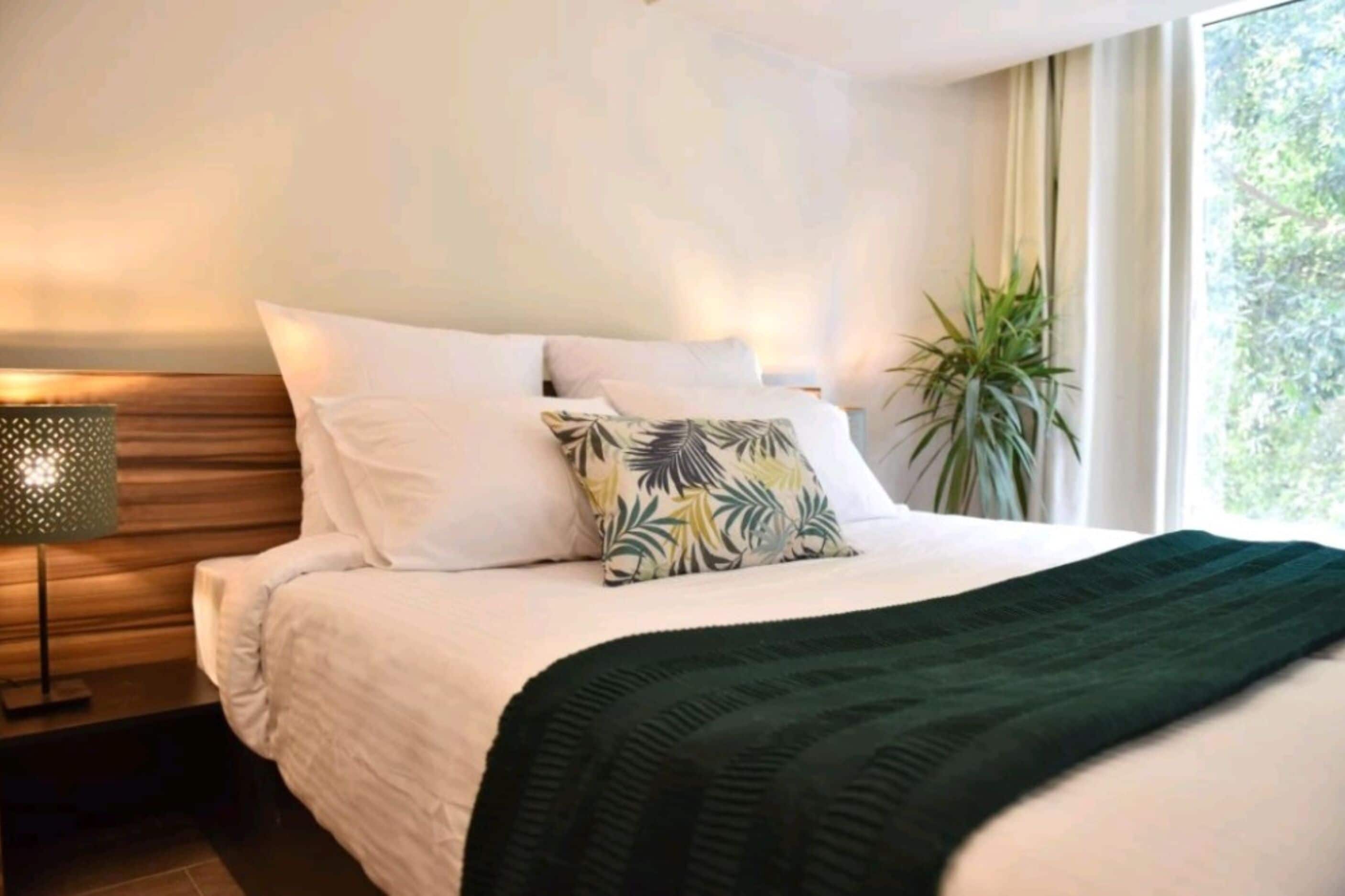Un lit bien fait, avec du linge de lit blanc et un plaid vert, une lampe de chevet et une plante en pot près de la fenêtre.