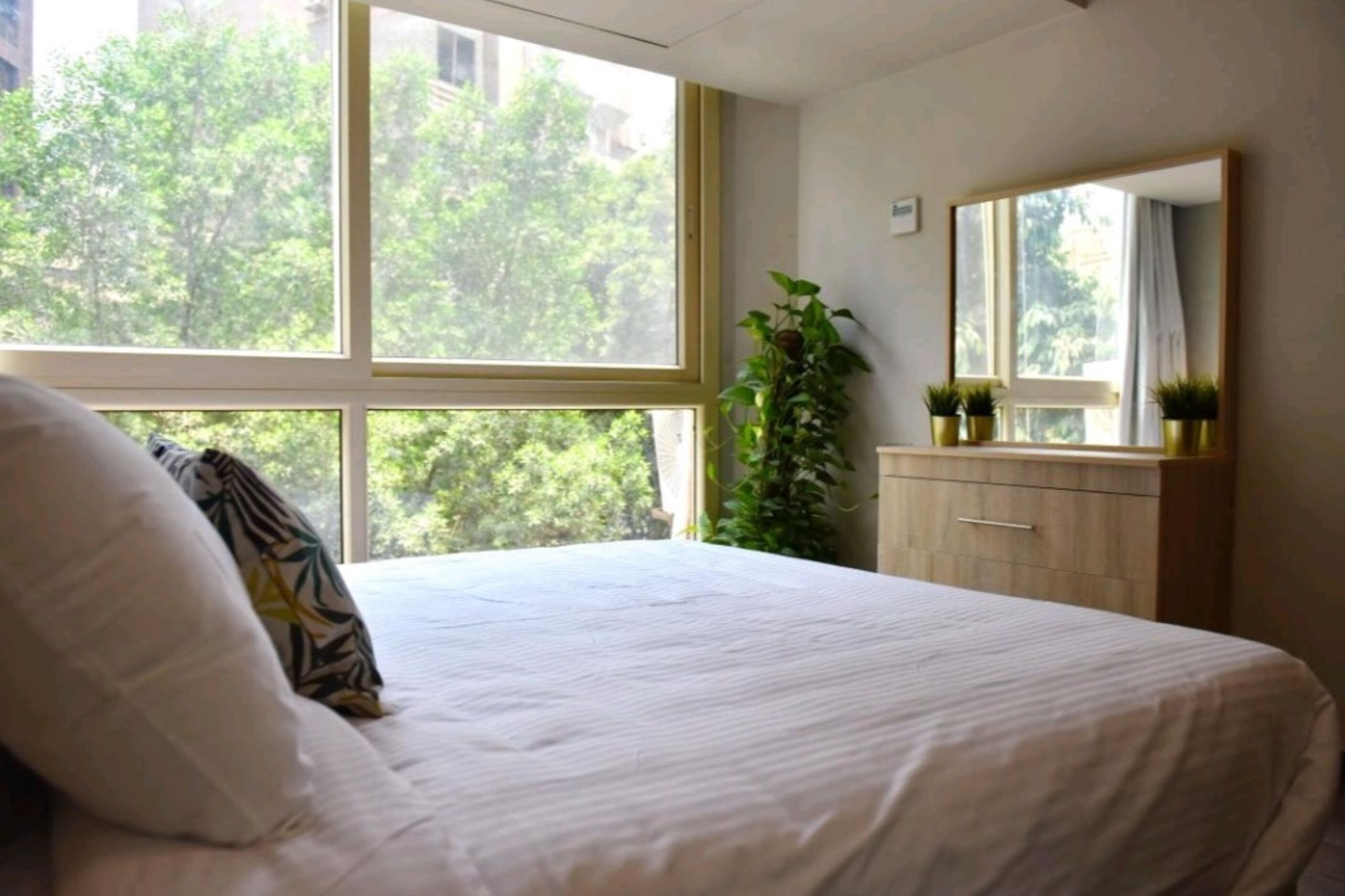 Un dormitorio con cama, cómoda, espejo y ventana con vista a árboles.