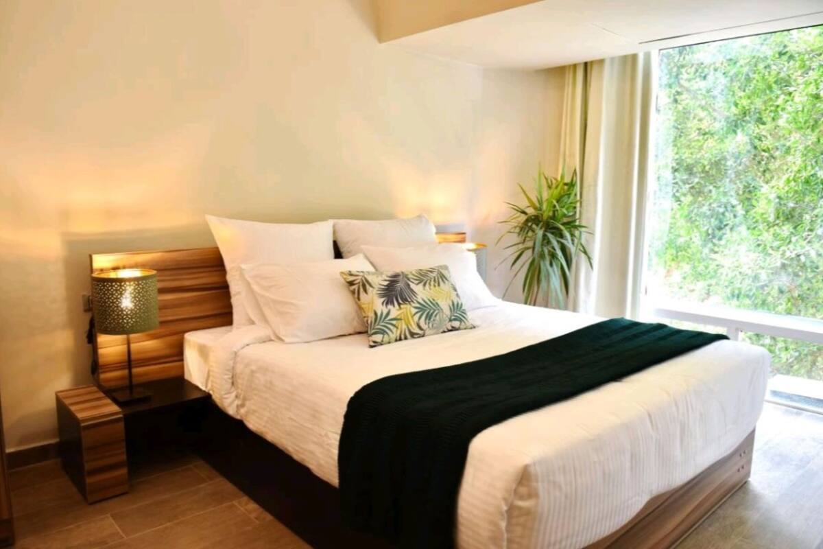 Un dormitorio con cama, mesitas de noche, lámpara, ventana con vista a vegetación y planta en maceta.