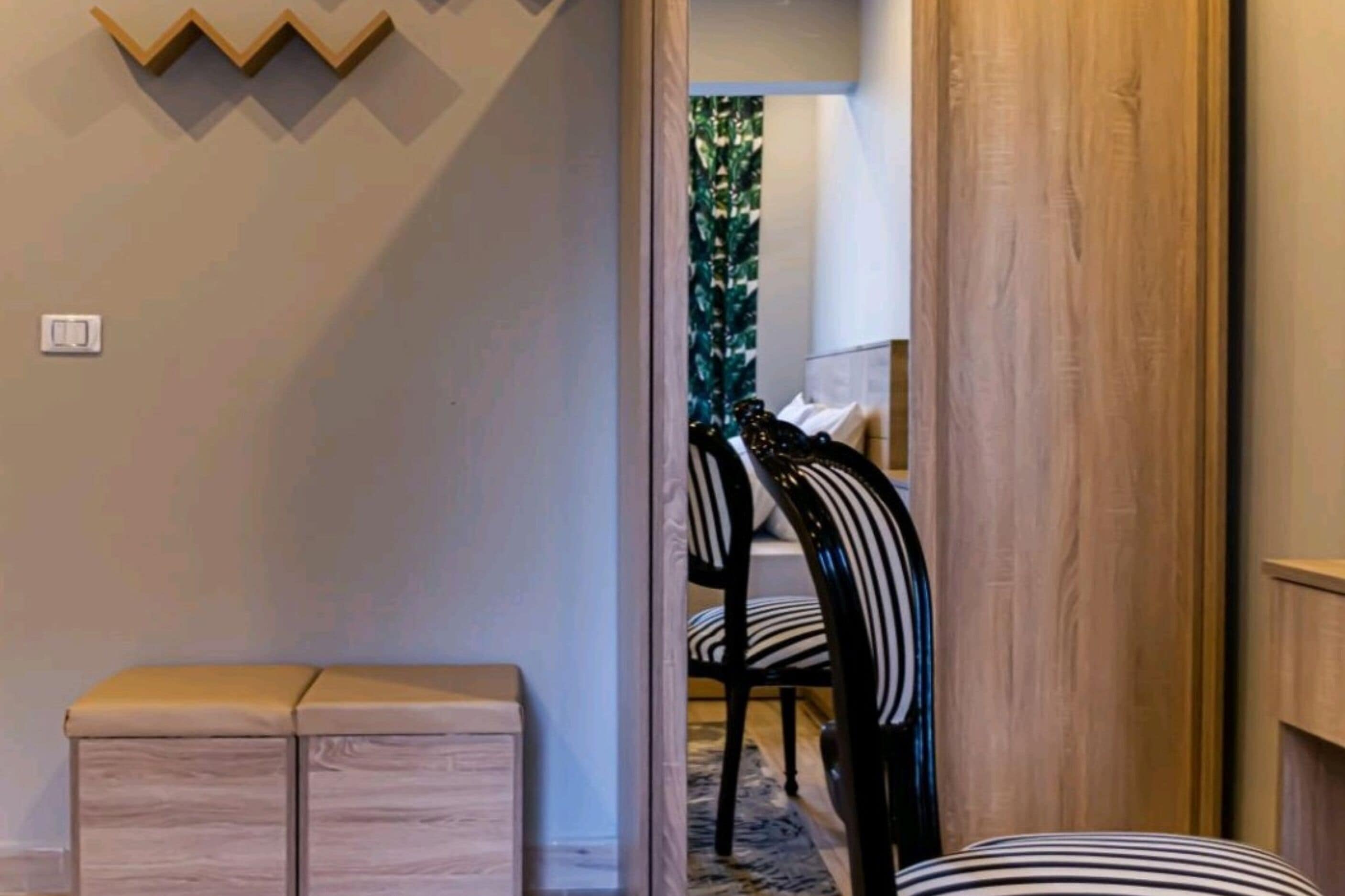 A hallway with a wooden bench, a wall-mounted light switch, and a door leading to a room with striped chairs.