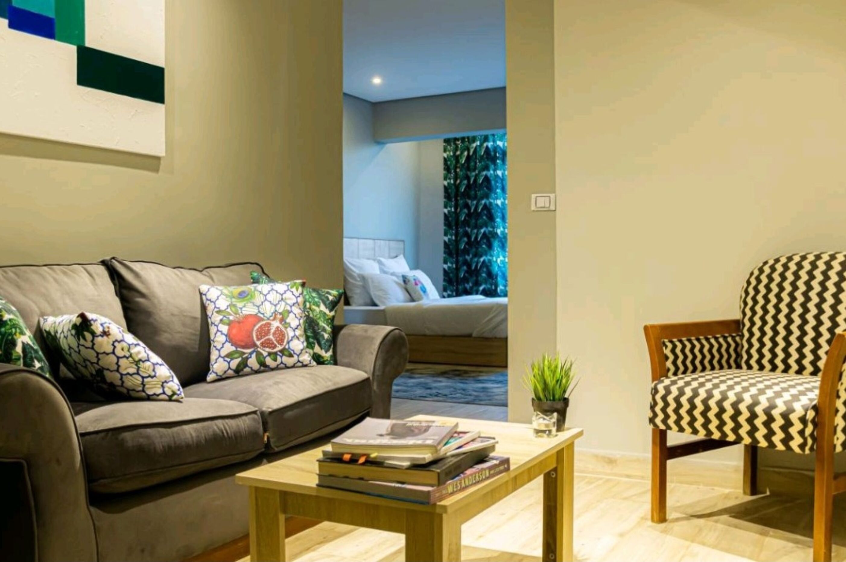 A living room with a sofa, a patterned armchair, a coffee table with books, and a view into a bedroom with a bed and a green wall.
