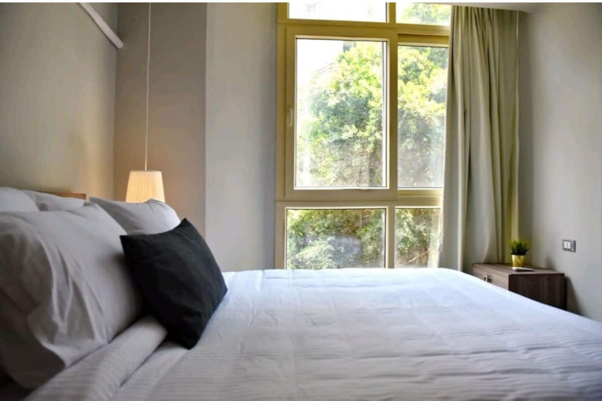 Un dormitorio con cama, lámpara de noche, ventana con cortinas y una pequeña mesa con una planta.