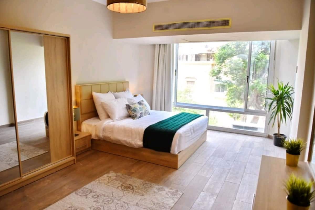 Un dormitorio con cama, espejo, ventana y una planta.