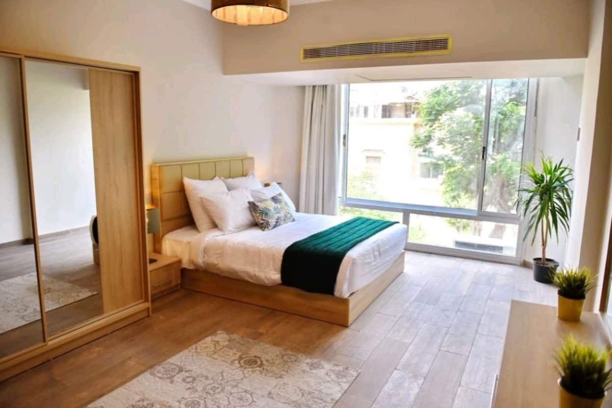 Un dormitorio con cama, espejo, ventana y una planta.