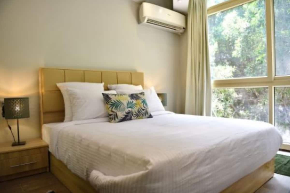 Un dormitorio con cama, mesita de noche, lámpara y ventana con vistas a árboles.