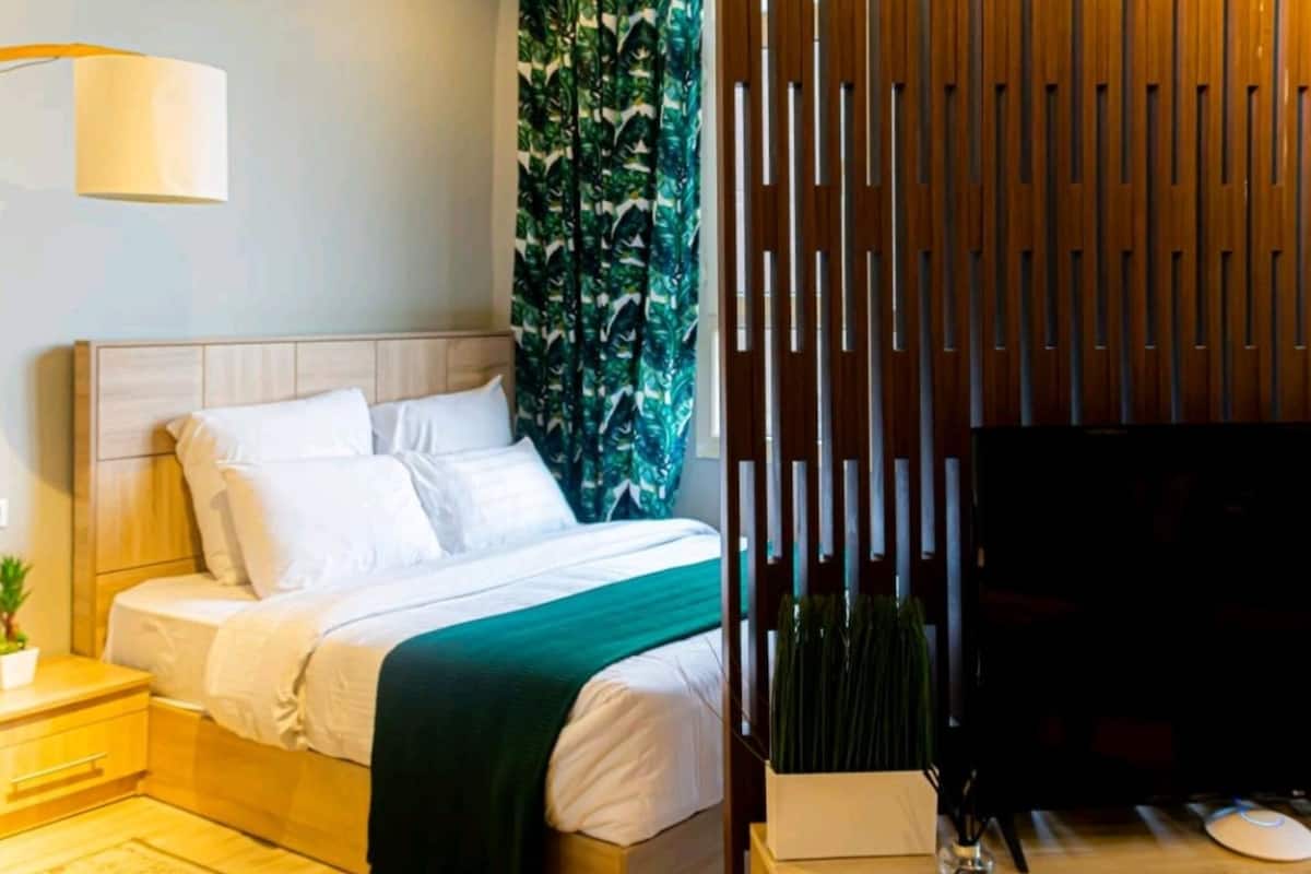 Un dormitorio con cama, mesita de noche, lámpara, televisor y un panel decorativo.