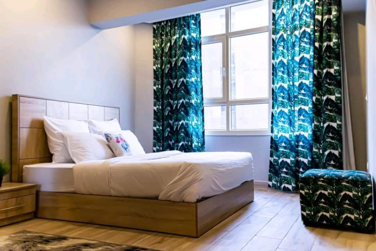 Un dormitorio con cama, mesita de noche, ventana con cortinas y un pequeño banco.