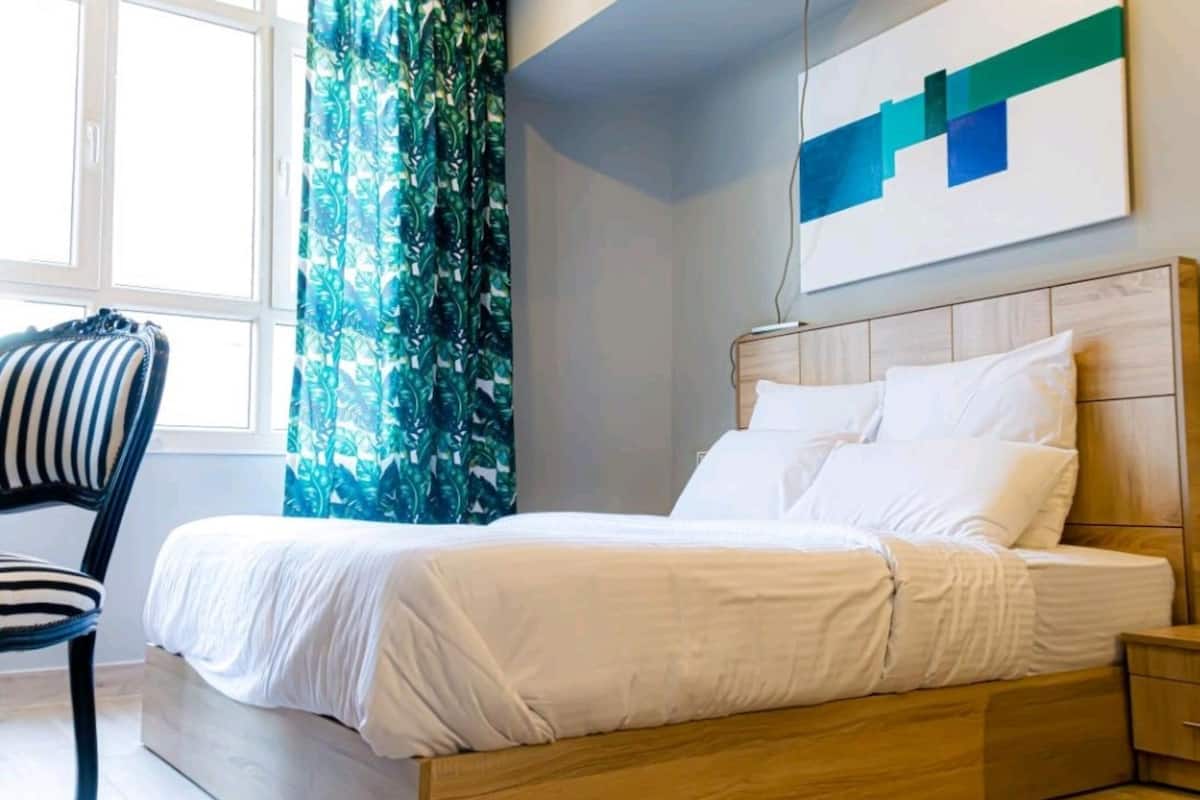 Una cama con ropa de cama blanca, cabecera de madera, mesita de noche, una silla y una ventana con cortinas.
